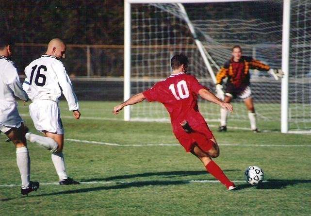 A student in a red uniform kicks towards the goal in a soccer game.