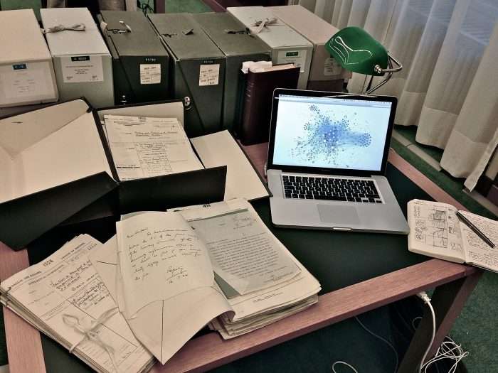A desk is covered in paperwork, file boxes, and a laptop computer.