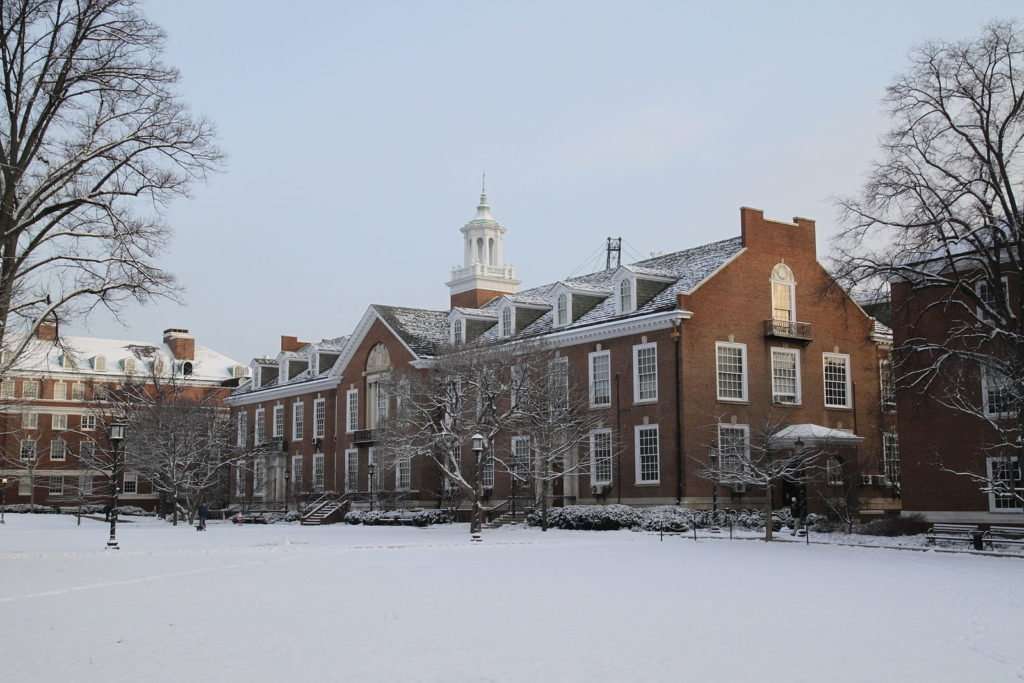A brick building with a steeple is featured beyond a lawn covered in snow at Johns Hopkins University.